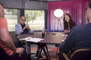 Vier Personen sitzen an einem runden Tisch in einem modernen Raum mit lila Vorhängen, diskutieren und schauen sich an.