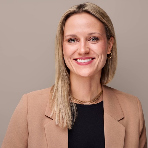 Porträt einer lächelnden Frau mit blonden Haaren, die einen beigen Blazer trägt, vor einem neutralen Hintergrund.
