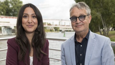 Zwei Personen sitzen am Ufer eines Gewässers, lächeln in die Kamera, umgeben von Bäumen und einer Brücke im Hintergrund.