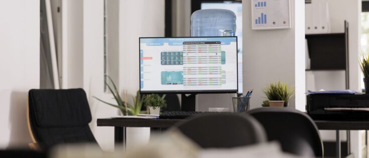Leerer Bürotisch mit Computerbildschirm, der Datenanalyse-Software und Finanzresultate zeigt. Pflanzen und Wasserbehälter im Hintergrund.