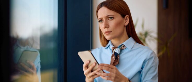Nachdenkliche Geschäftsfrau in blauem Hemd, hält Smartphone und blickt besorgt aus dem Bürofenster.