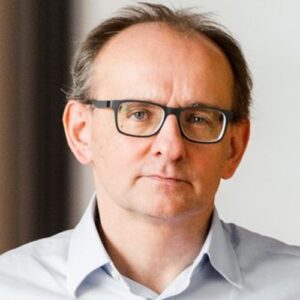 Porträt eines Mannes mit Brille, kurzen Haaren und ernstem Gesichtsausdruck, in einem hellen Raum.