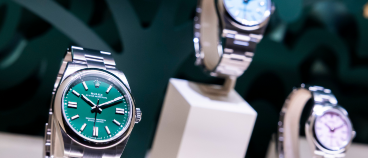 Drei Rolex-Uhren auf Ständern, eine mit grünem Ziffernblatt, zwei weitere mit blauen und lila Ziffernblättern.