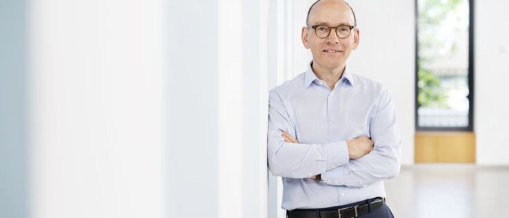Martin Roth, älterer Herr mit Brille, hellblaues Hemd, steht mit verschränkten Armen in einem hellen Flur und lächelt.