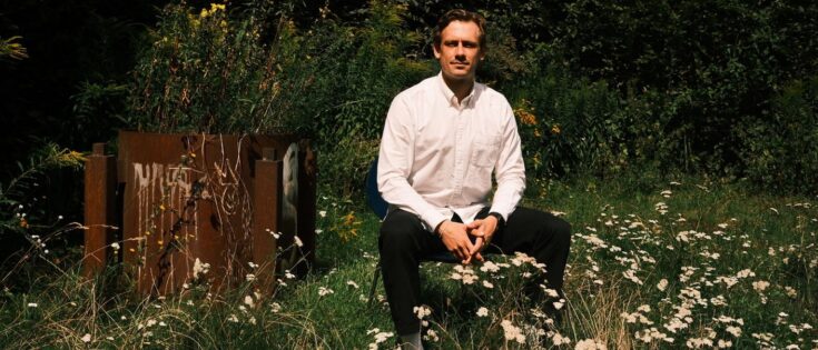 Mann in weißem Hemd sitzt auf einer Wiese mit Blumen, hinter ihm eine rustikale Holzstruktur und grüne Pflanzen.