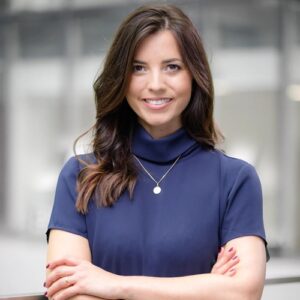 Frau mit langen, braunen Haaren in blauer Bluse, lächelt und steht mit verschränkten Armen vor einem modernen Hintergrund.