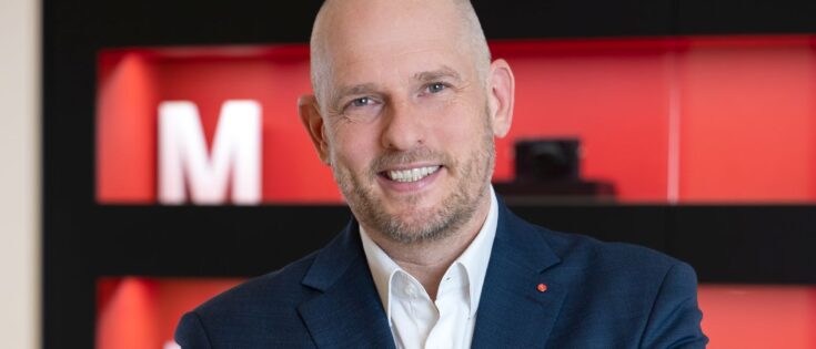 Mann in Anzug lächelt vor einem modernen Hintergrund mit roten und schwarzen Elementen.